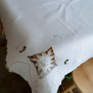 May include: White linen tablecloth with delicate embroidered floral designs. The tablecloth has a scalloped edge and is perfect for a formal dinner party.