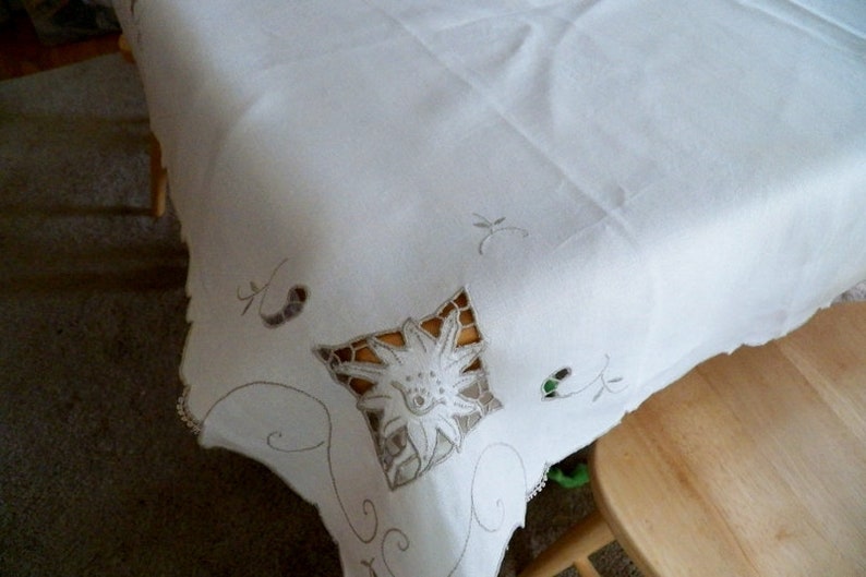 May include: White linen tablecloth with delicate embroidered floral designs. The tablecloth has a scalloped edge and is perfect for a formal dinner party.