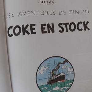 May include: A book page featuring the title Les Aventures de Tintin and Coke en Stock in black text, with the author name Herg&eacute; above. Below the text is a circular illustration of a ship at sea viewed through a crosshair scope sight.