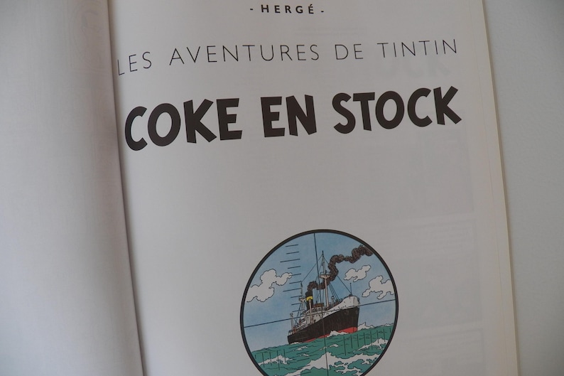 May include: A book page featuring the title Les Aventures de Tintin and Coke en Stock in black text, with the author name Herg&eacute; above. Below the text is a circular illustration of a ship at sea viewed through a crosshair scope sight.
