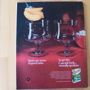 May include: An advertisement featuring two clear stemmed glasses with a peach and water droplets above them. The text reads "Spots are never in good taste" and "So get the Cascade look... virtually spotless."