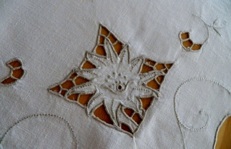 May include: White linen tablecloth with delicate embroidered floral designs. The tablecloth features a large, intricate flower in the center and smaller floral motifs around the edges.