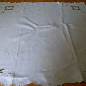 May include: White linen tablecloth with delicate embroidered floral designs in a brown thread. The tablecloth has a scalloped edge.