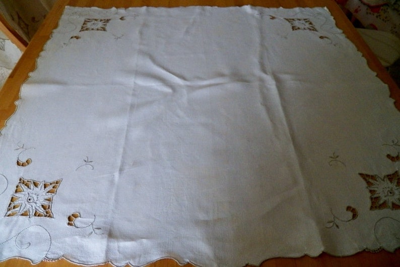May include: White linen tablecloth with delicate embroidered floral designs in a brown thread. The tablecloth has a scalloped edge.