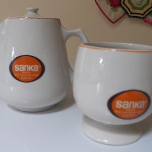 Puede incluir: Una tetera y una taza de cerámica blanca con un logotipo naranja que dice "Sanka Brand 99% Caffeine Free Coffee".