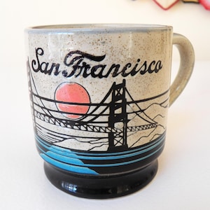 May include: A ceramic mug with a beige speckled finish, featuring a black and blue illustration of the Golden Gate Bridge, a red sun, and the words "San Francisco". The mug has a black base and a curved handle.
