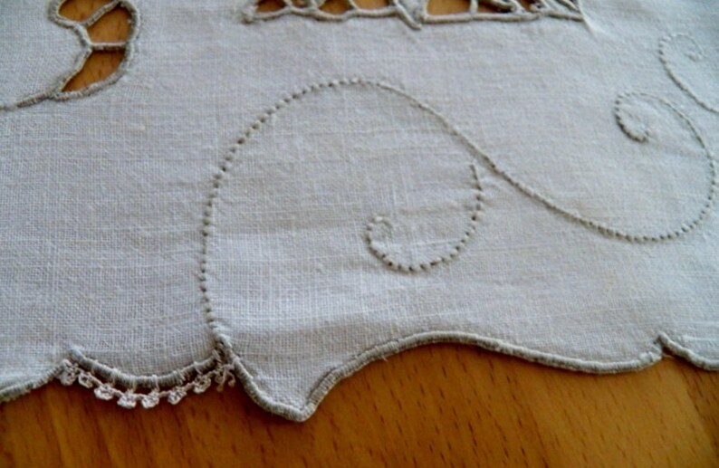 May include: A white linen tablecloth with a scalloped edge and delicate embroidered details. The embroidery features a repeating pattern of swirls and loops.