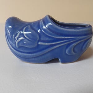 May include: A blue ceramic clog-shaped planter with a floral design. The planter is a vibrant blue colour and has a glossy finish. The floral design is in white and is on the side of the clog. The planter is a decorative item.