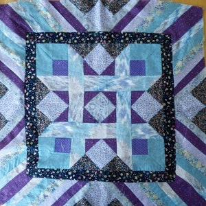 May include: A handmade quilt with a geometric design. The quilt features a central square with intersecting light blue and purple shapes, surrounded by a border of floral fabric and diagonal stripes in shades of blue and purple.