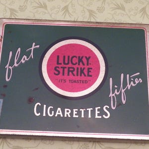 May include: A vintage tin box for Lucky Strike cigarettes. The box is green with a red and white circle in the center that says "Lucky Strike" and "It's Toasted". The words "flat fifties" are written diagonally across the box.