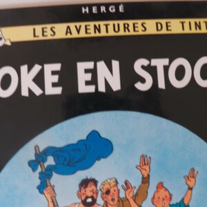 May include: A hardcover book titled Coke en Stock from the Les Aventures de Tintin series by Herg&eacute;. The black cover features a circular illustration of three characters waving, with a small graphic of Tintin and Snowy in the top left corner.