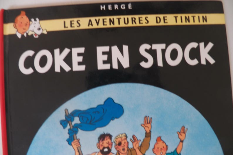 May include: A hardcover book titled Coke en Stock from the Les Aventures de Tintin series by Herg&eacute;. The black cover features a circular illustration of three characters waving, with a small graphic of Tintin and Snowy in the top left corner.