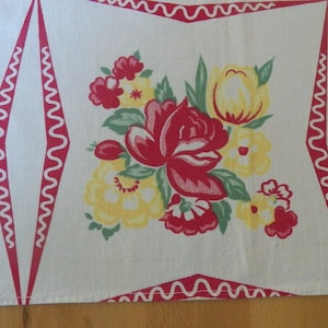May include: A white fabric runner featuring a central floral bouquet of red and yellow flowers with green leaves. The design is framed by red diamond-shaped borders with white wavy lines, and partial fruit illustrations are visible on the sides.