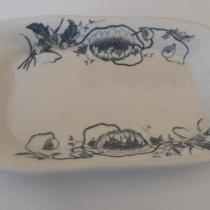 Puede incluir: Una bandeja rectangular de cerámica blanquecina con un diseño floral en azul oscuro. El diseño presenta flores y hojas estilizadas en los bordes. La bandeja tiene una forma ligeramente curvada.