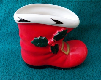 May include: A ceramic Christmas boot planter, painted red with a white rim and black accents. The boot features a green holly and berry design and a gold-colored buckle. This festive planter is designed for holiday decor.