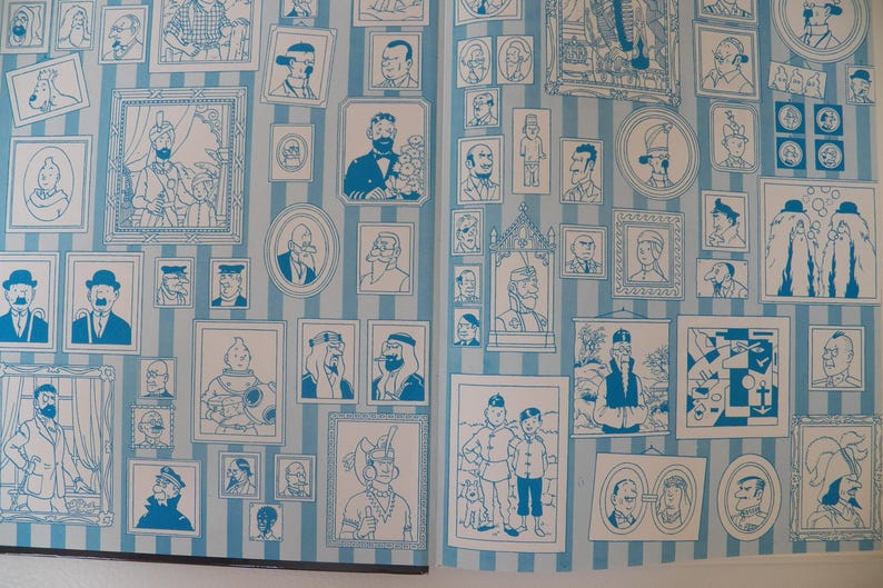 May include: A light blue and white vertical striped wallpaper featuring a repeating pattern of various framed illustrations of characters and scenes in blue ink. The design includes numerous portraits, rectangular frames, and oval frames.