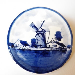 May include: A round, blue and white ceramic plate featuring a windmill scene. The design depicts a windmill, houses, and trees in a rural landscape. The plate has a blue border and is likely decorative.