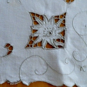May include: White linen tablecloth with delicate embroidered floral designs. The tablecloth features a central flower motif with intricate cutouts and a scalloped edge.