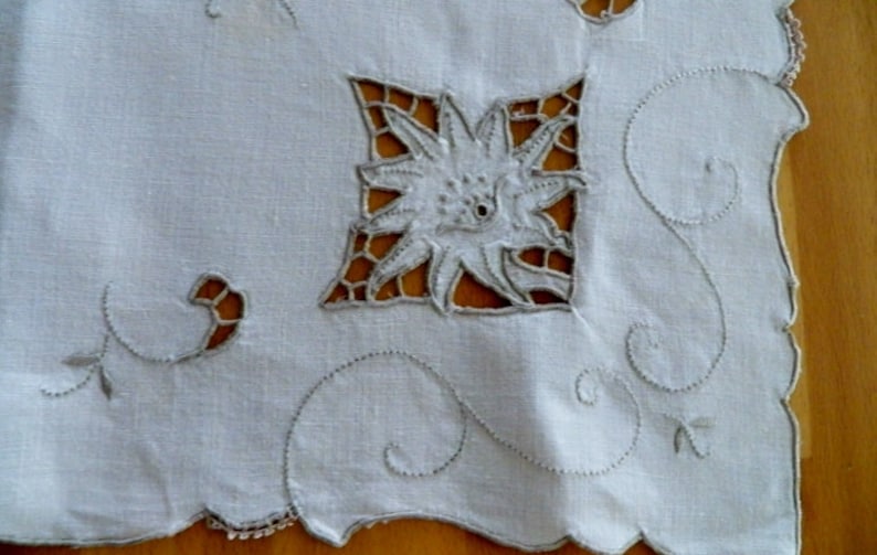 May include: White linen tablecloth with delicate embroidered floral designs. The tablecloth features a central flower motif with intricate cutouts and a scalloped edge.