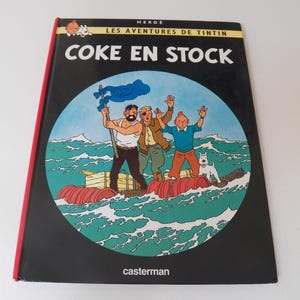 May include: A hardcover book titled Les Aventures de Tintin: Coke en Stock by Herg&eacute;. The black cover features a circular illustration of three characters and a white dog on a makeshift raft made of red barrels and wooden crates in a green sea.