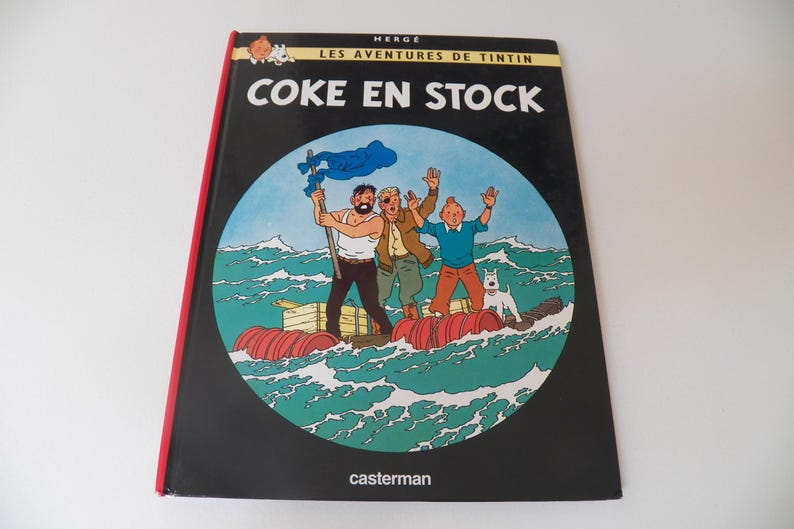 May include: A hardcover book titled Les Aventures de Tintin: Coke en Stock by Herg&eacute;. The black cover features a circular illustration of three characters and a white dog on a makeshift raft made of red barrels and wooden crates in a green sea.