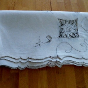 May include: A white linen tablecloth with a scalloped edge and delicate embroidery. The embroidery features a floral design with a central flower and scrolling vines.