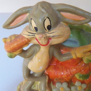 Puede incluir: Figura de cerámica vintage de Bugs Bunny sosteniendo una zanahoria. La figura está pintada en tonos de gris, naranja, verde y amarillo. Bugs Bunny sonríe con la boca roja. Artículo decorativo.
