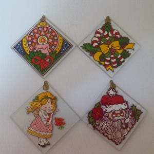 May include: Four diamond-shaped glass ornaments featuring colorful holiday illustrations. One shows a lit candle with holly, another candy canes with a yellow bow, one a blonde figure in a pink dress, and the last a Santa Claus face with a red hat.
