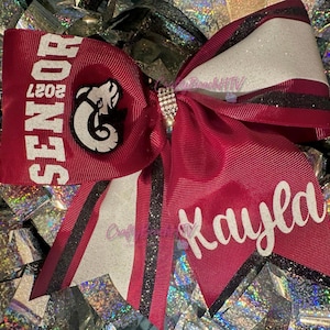 May include: A maroon cheer bow featuring the text SENIOR 2027 and a ram mascot graphic on one side, with the name Kayla in white script on the other. The bow includes black glitter accents, white ribbon layers, and a central rhinestone knot.