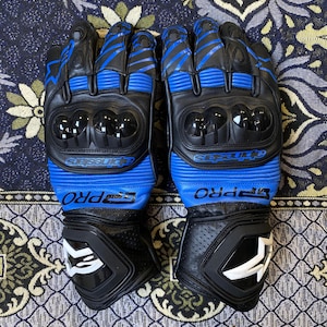 Pode incluir: Um par de luvas de motociclismo pretas e azuis com o texto "SP PRO" e "Alpinestars". As luvas têm proteção para os nós dos dedos e detalhes brancos. As luvas são de couro.