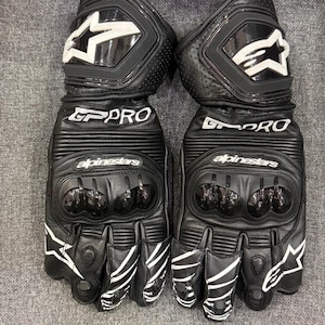 Pode incluir: Par de luvas de motociclismo em couro preto com detalhes brancos. As luvas apresentam a inscrição "GP PRO" e a marca "Alpinestars". Possuem proteção para os nós dos dedos e um logotipo de estrela no punho. Projetadas para motociclismo.