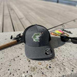 Puede incluir: Una gorra de béisbol gris carbón con parte trasera de malla negra y un gráfico de pez verde bordado en el frente. La gorra presenta una pegatina circular en la visera con el texto 110 y Flexfit, una corona estructurada y visera curva.