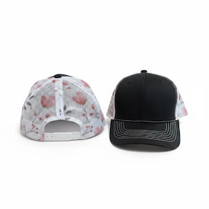 May include: Two baseball caps are shown. One cap is black with a solid front panel and a white mesh back featuring a red floral pattern. The second cap is black with a solid front panel and a white mesh back, featuring a snapback closure.