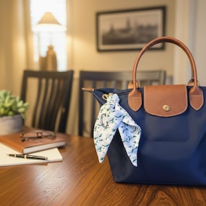 Op de afbeelding: Een marineblauwe nylon handtas met bruine lederen handvatten en een bijpassende bruine lederen klep met een goudkleurige drukknoopsluiting. Een witte zijden sjaal met een groen bloemenpatroon en een zwarte geborduurde letter C is aan het handvat geknoopt.