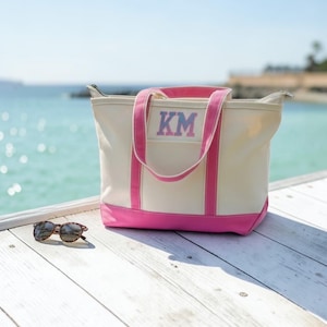 Puede incluir: Un bolso de lona color crema con ribetes y asas de color rosa brillante. La parte frontal del bolso presenta las letras KM bordadas en una fuente de bloque. Junto al bolso hay un par de gafas de sol con montura marrón.