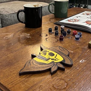 May include: A tattoo parlor interior with framed tattoo art on the brick wall. A wooden table holds a skull-shaped decoration, ink bottles, a lighter, and a tattoo design book. Two mugs sit on the table, and a sofa is in the background.