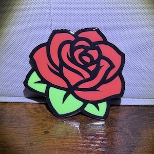 May include: A red rose-shaped pin with black outlines and green leaves. The pin is a vibrant, colorful accessory, perfect for adding a touch of floral flair to clothing or accessories. The design is bold and eye-catching.