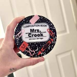 Op de afbeelding: Een ronde onderzetter met een ontwerp van een compositieboek. De onderzetter heeft de tekst "COMPOSITION BOOK" en "Mrs. Crook". Het bevat illustraties van een paperclip, bindclip, kleurpotlood en gum. De achtergrond is zwart met witte spikkels.