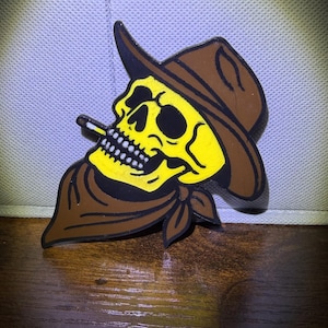 May include: A yellow skull wearing a brown cowboy hat and bandana, with a lit cigarette in its mouth. The skull has white teeth and black outlines, set against a dark brown and yellow color scheme.