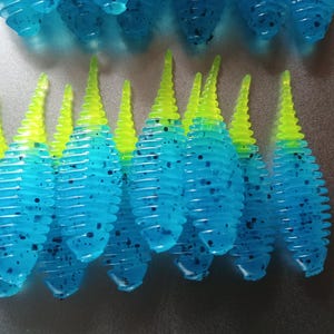 May include: A collection of soft plastic fishing lures featuring a ribbed, translucent blue body with black speckles and a bright neon yellow tapered tail. The lures are arranged in rows, showcasing their segmented design and vibrant color contrast.