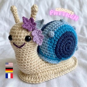 May include: A beige crocheted snail toy with a blue and dark blue spiral shell, black plastic eyes, and a purple crocheted flower accent. The text CROCHET PATTREN appears in pink letters above the snail, along with small US, German, and French flags.