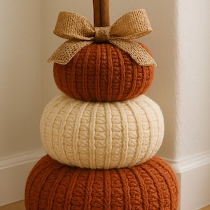 May include: A stacked pumpkin decoration made of three crocheted spheres in alternating rust orange and cream colors. The top features a brown cinnamon stick stem and a tan burlap ribbon bow. A circular graphic includes the text PDF Pattern.