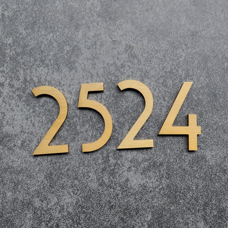 May include: Four individual house numbers forming the sequence 2524, crafted from a brushed gold-toned metal with a smooth, matte finish. Each digit features a clean, sans-serif typeface with sharp, angular edges and a consistent metallic texture.