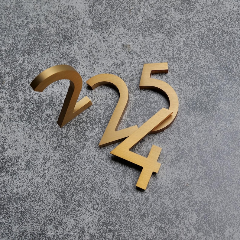 May include: Three metallic gold house numbers, specifically the digits 2, 2, and 5, arranged in a cluster. Each number features a clean, sans-serif typeface with a brushed finish, creating a reflective surface that catches light across the edges.