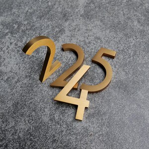 May include: Three dimensional house numbers 2, 2, and 5 crafted from brushed gold metal. The numbers feature a clean, sans-serif font with a smooth, metallic finish. Each digit is positioned to overlap slightly, creating a layered arrangement.