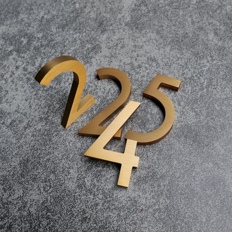 May include: Three dimensional house numbers 2, 2, and 5 crafted from brushed gold metal. The numbers feature a clean, sans-serif font with a smooth, metallic finish. Each digit is positioned to overlap slightly, creating a layered arrangement.