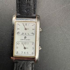 Puede incluir: Un reloj de pulsera rectangular Pierre Cardin con caja plateada y correa de cuero negro. La esfera del reloj tiene dos esferas separadas, una con n&uacute;meros romanos y otra con n&uacute;meros ar&aacute;bigos. El reloj muestra la hora.