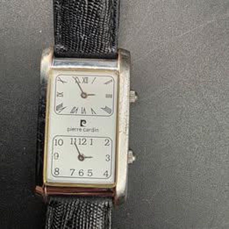 Puede incluir: Un reloj de pulsera rectangular Pierre Cardin con caja plateada y correa de cuero negro. La esfera del reloj tiene dos esferas separadas, una con n&uacute;meros romanos y otra con n&uacute;meros ar&aacute;bigos. El reloj muestra la hora.