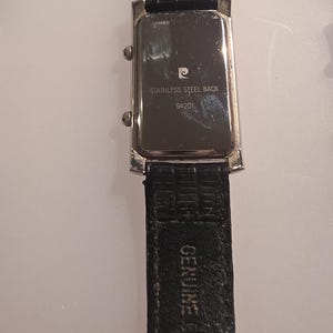 Puede incluir: Vista trasera de un reloj de pulsera rectangular con una correa de cuero negro. La esfera del reloj es plateada con el texto "STAINLESS STEEL BACK" y "94201". La correa est&aacute; marcada con "GENUINE LEATHER".