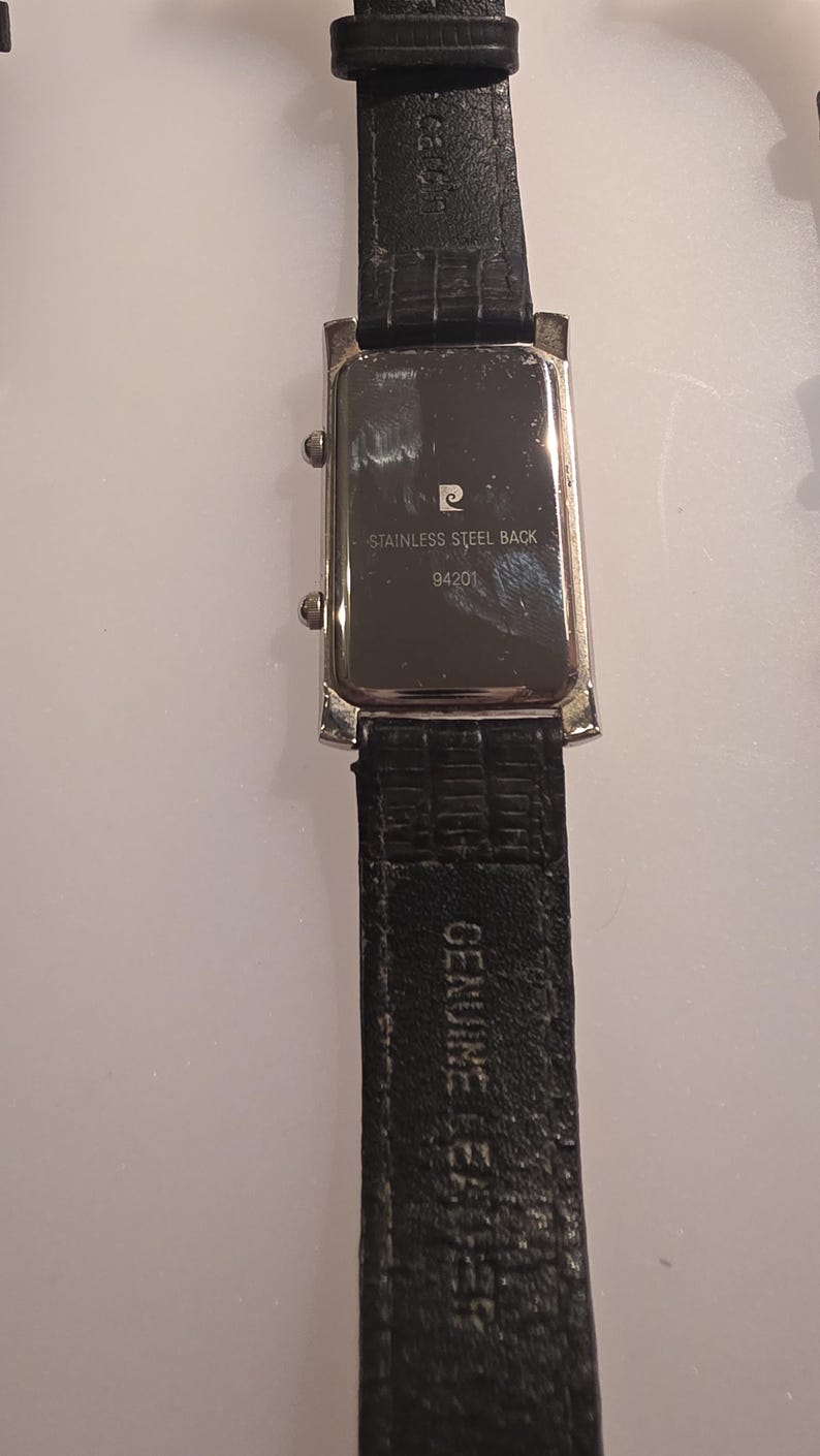 Puede incluir: Vista trasera de un reloj de pulsera rectangular con una correa de cuero negro. La esfera del reloj es plateada con el texto "STAINLESS STEEL BACK" y "94201". La correa est&aacute; marcada con "GENUINE LEATHER".
